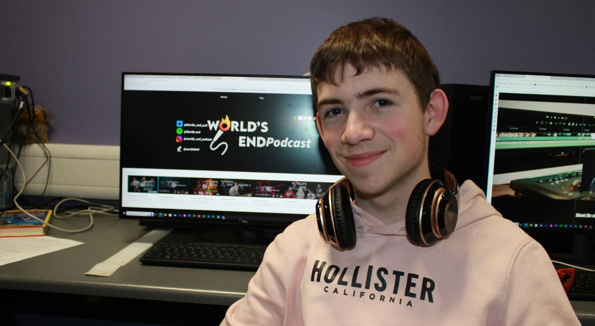 Young man with short dark hear with headphones around his neck smiling to camera with computer screens in background featuring World's End Podcast logo